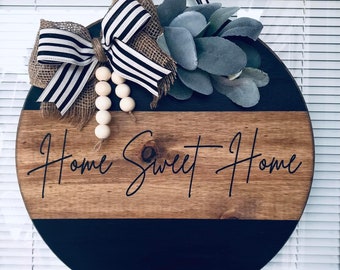 May include: A round wooden door hanger with a black and brown wood design. The top half of the hanger is brown wood with the words "Home Sweet Home" written in black script. The bottom half of the hanger is black. The hanger is decorated with a burlap bow with black and white stripes, a cluster of faux succulents, and a strand of wooden beads.