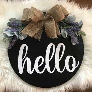 Op de afbeelding: Een rond, zwart houten bord met het woord "hello" in witte, cursieve letters. Het bord is versierd met een strik van jute en takjes lavendel.