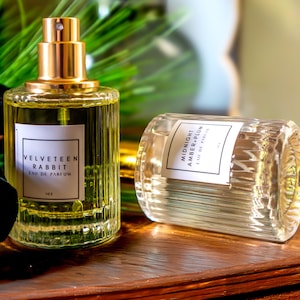 May include: Two cylindrical glass perfume bottles with vertical ribbing. One bottle stands upright with a gold spray nozzle and a label reading Velveteen Rabbit Eau de Parfum 1 oz. The other lies horizontally with a label reading Midnight Amber+Plum 1 oz.