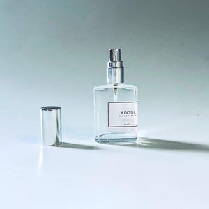 May include: A clear glass perfume bottle with a silver spray top and a matching cap. The bottle has a rectangular shape and a white label with the word "WOODS" printed on it. The bottle contains 15 ml of perfume.