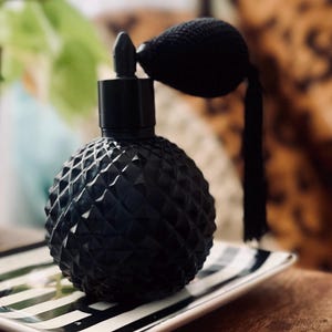 May include: A black glass perfume bottle with a diamond-shaped pattern and a black bulb atomizer. The bottle is sitting on a black and white striped tray.