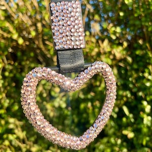 May include: A heart-shaped accessory with a black strap and a rectangular clasp, both adorned with sparkling, light pink rhinestones. The heart is outlined with the same rhinestones, creating a glamorous and eye-catching design. The background is a blurred view of green foliage.