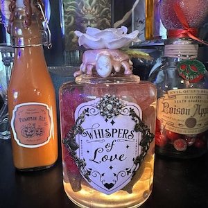 May include: A collection of decorative glass bottles with unique designs. One bottle features a label that reads "Whispers of Love" and is adorned with a decorative frame. Another bottle has a "Pumpkin Ale" label. The bottles are lit from within, creating a warm glow.