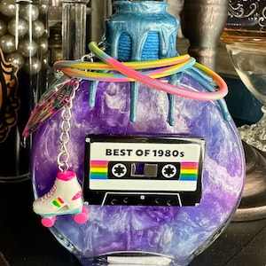 Puede incluir: Una botella decorativa redonda, morada, con una parte superior azul y detalles goteantes. Un dije de cinta de cassette con el texto "BEST OF 1980s" est&aacute; adjunto, junto con un dije de pat&iacute;n y un cord&oacute;n arco&iacute;ris.