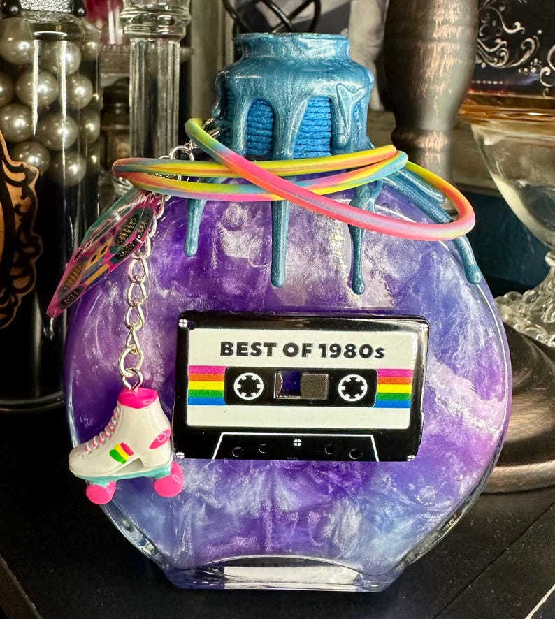 Puede incluir: Una botella decorativa redonda, morada, con una parte superior azul y detalles goteantes. Un dije de cinta de cassette con el texto "BEST OF 1980s" est&aacute; adjunto, junto con un dije de pat&iacute;n y un cord&oacute;n arco&iacute;ris.