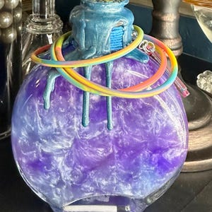 Puede incluir: Una botella de vidrio redonda llena de un l&iacute;quido morado arremolinado. La botella tiene una parte superior azul texturizada con detalles goteados. Una pulsera de arco&iacute;ris envuelve el cuello. Hay una peque&ntilde;a etiqueta rectangular en la parte inferior.