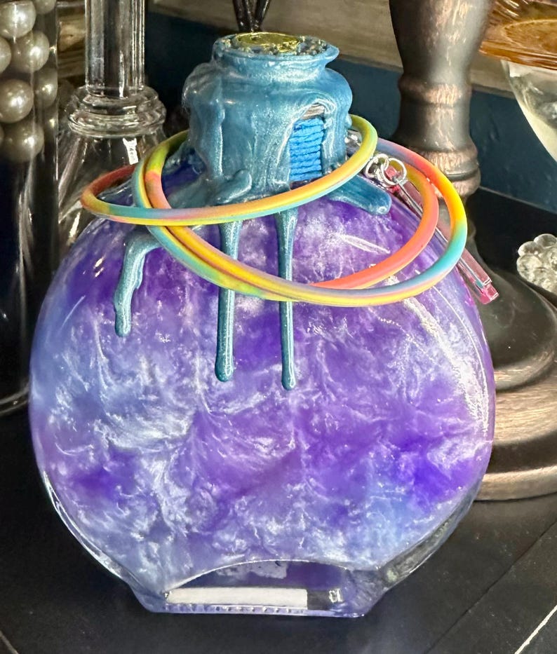 Puede incluir: Una botella de vidrio redonda llena de un l&iacute;quido morado arremolinado. La botella tiene una parte superior azul texturizada con detalles goteados. Una pulsera de arco&iacute;ris envuelve el cuello. Hay una peque&ntilde;a etiqueta rectangular en la parte inferior.