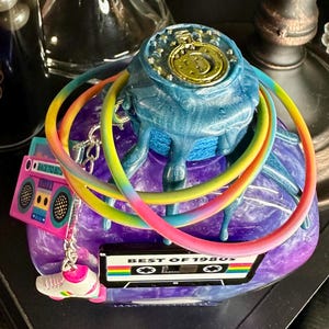 Puede incluir: Una escultura decorativa con un tema retro de los a&ntilde;os 80. La escultura presenta una base azul y morada con un dise&ntilde;o de reloj dorado en la parte superior. Se adjuntan anillos de colores arco&iacute;ris y dijes de una radio, patines y una cinta de cassette.