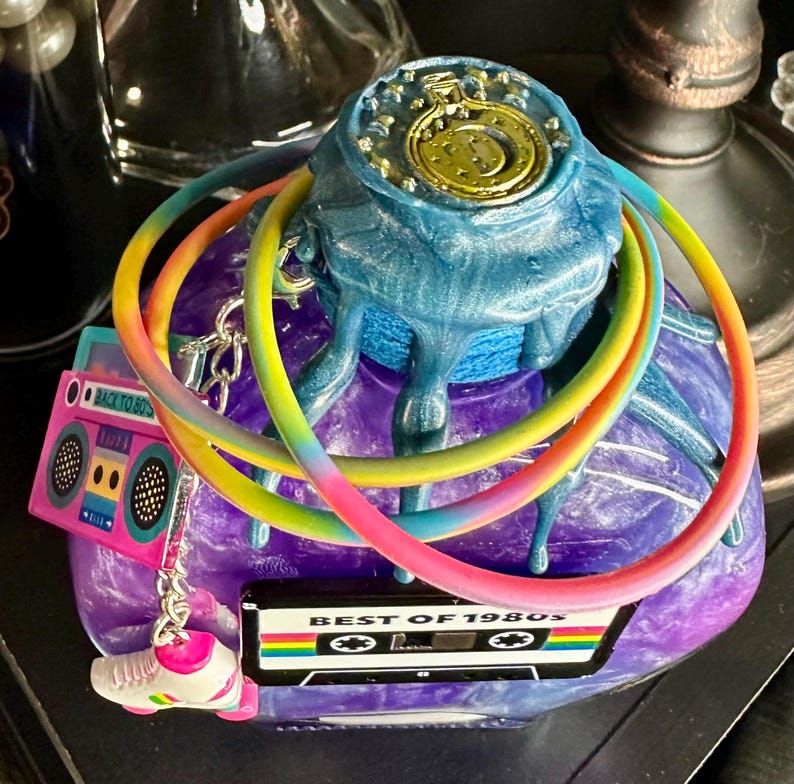 Puede incluir: Una escultura decorativa con un tema retro de los a&ntilde;os 80. La escultura presenta una base azul y morada con un dise&ntilde;o de reloj dorado en la parte superior. Se adjuntan anillos de colores arco&iacute;ris y dijes de una radio, patines y una cinta de cassette.