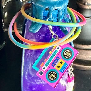 Puede incluir: Una botella morada con una tapa azul y un cord&oacute;n arco&iacute;ris enrollado alrededor. Un dije de cinta de cassette rosa con el texto "BACK TO 80'S" cuelga de la botella. La botella est&aacute; decorada con un dise&ntilde;o dorado en la parte superior.