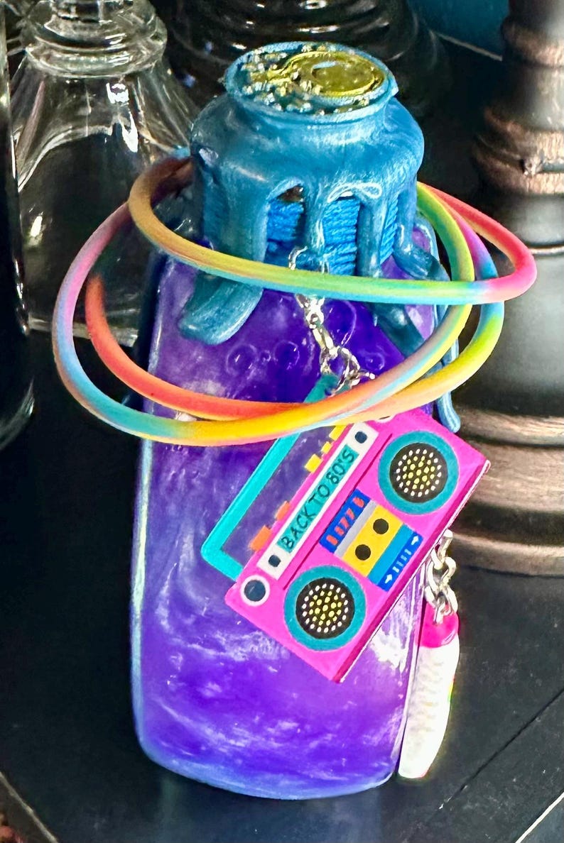Puede incluir: Una botella morada con una tapa azul y un cord&oacute;n arco&iacute;ris enrollado alrededor. Un dije de cinta de cassette rosa con el texto "BACK TO 80'S" cuelga de la botella. La botella est&aacute; decorada con un dise&ntilde;o dorado en la parte superior.