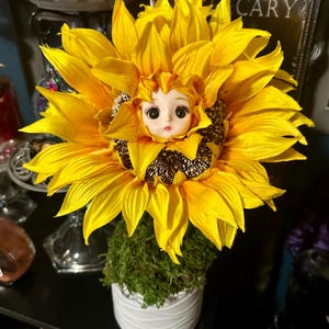 May include: A decorative sunflower ornament featuring a doll face at its center. The large yellow fabric petals surround the face, which has dark eyes and pink lips. The flower sits atop a green moss-covered base inside a white cylindrical pot.