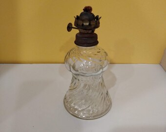 May include: A clear glass oil lamp with a twisted design. The lamp has a metal top with a wick holder and a small, round knob.
