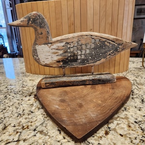 May include: A weathered, hand-carved wooden duck sculpture with a distressed paint finish in shades of white, black, and brown. The duck is mounted on a heart-shaped wooden base, creating a rustic, vintage aesthetic. The sculpture is a decorative item.