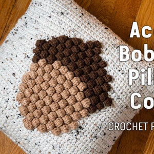 May include: A white crocheted pillow cover with a brown and beige acorn design. The pillow cover features a bobble stitch pattern. The text "Acorn Bobble Pillow Cover CROCHET PATTERN" is displayed on the right side of the image.