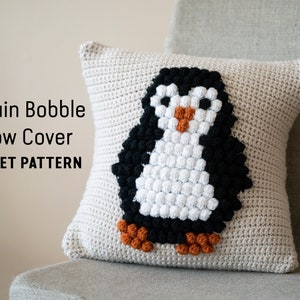 May include: A beige crocheted pillow cover with a penguin design. The penguin is made of black, white, and orange bobbles. The pillow cover has the text "Penguin Bobble Pillow Cover CROCHET PATTERN" on it.