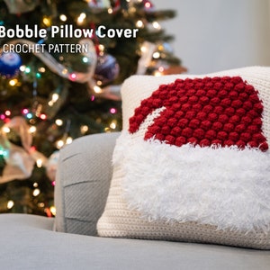 May include: A crocheted pillow cover with a Santa hat design. The hat is red with white trim and has a white fluffy pom-pom. The pillow cover is beige and has a textured surface. "Santa Bobble Pillow Cover CROCHET PATTERN" is written in black text at the top of the image.