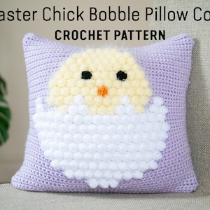 May include: Crochet pattern for an Easter chick bobble pillow cover. The pillow cover is purple with a white and yellow chick design. The chick is peeking out of a white egg.
