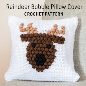 May include: White crochet pillow cover with a brown and beige reindeer face design. The reindeer has bobble nose and antlers.  Reindeer Bobble Pillow Cover CROCHET PATTERN