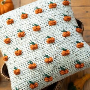 Könnte beinhalten: Ein quadratischer Kissenbezug aus Häkelarbeit in hellem Cremeweiß mit gesprenkelter Textur, der ein wiederkehrendes Muster aus kleinen, erhabenen orangefarbenen Kürbissen mit grünen Stielen auf der Vorderseite aufweist.