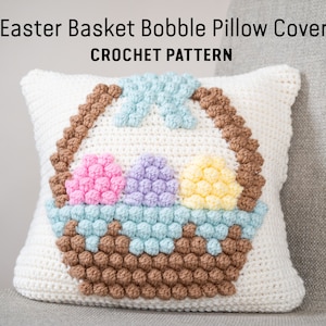 May include: Crochet pattern for an Easter basket cushion cover. The cushion cover features a white background with a brown basket filled with three pastel coloured eggs. The basket is made of brown bobbles and the eggs are made of pink, purple, and yellow bobbles.