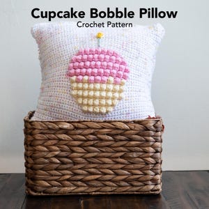 Può includere: Un cuscino bianco all'uncinetto con un design a cupcake rosa e giallo. Il cuscino è seduto in un cesto di vimini marrone. Il testo "Cupcake Bobble Pillow" e "Crochet Pattern" è sopra il cuscino.