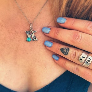 Puede incluir: Collar con colgante de plata con un colgante con la letra "K". El colgante presenta una piedra turquesa y un dise&ntilde;o del suroeste con motivos florales y de cactus. El collar lo lleva una persona con esmalte de u&ntilde;as azul y anillos de plata.