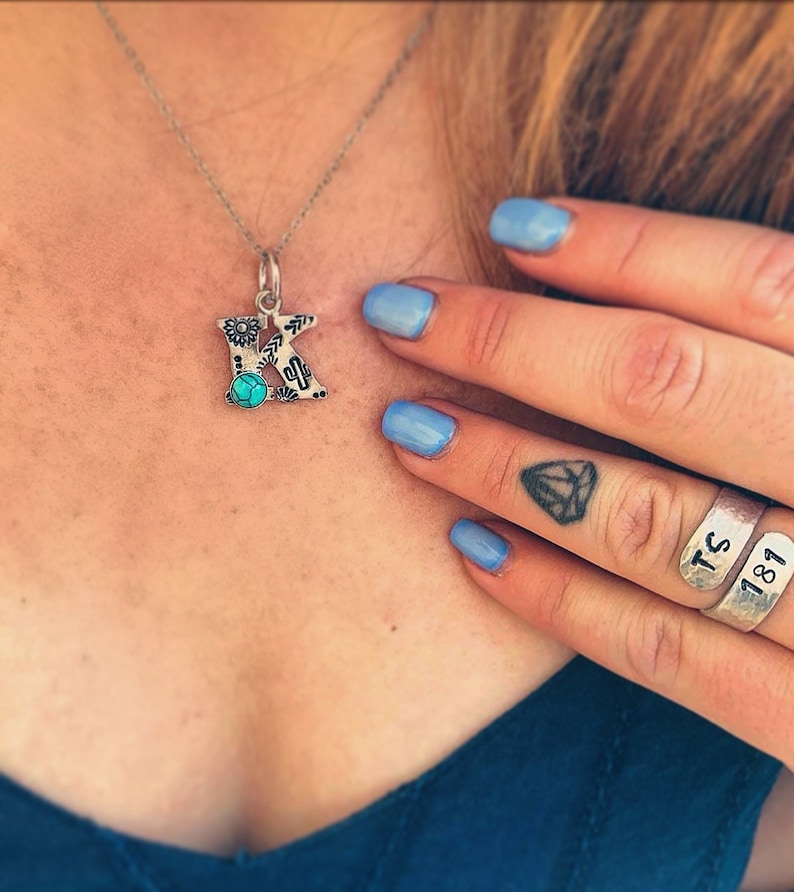 Puede incluir: Collar con colgante de plata con un colgante con la letra "K". El colgante presenta una piedra turquesa y un dise&ntilde;o del suroeste con motivos florales y de cactus. El collar lo lleva una persona con esmalte de u&ntilde;as azul y anillos de plata.