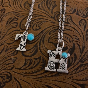Puede incluir: Dos collares con iniciales plateadas. La letra "T" presenta un dise&ntilde;o de cactus, y la letra "H" tiene un girasol. Ambos colgantes tienen una peque&ntilde;a piedra turquesa y cuelgan de delicadas cadenas. El fondo es una superficie marr&oacute;n texturizada.