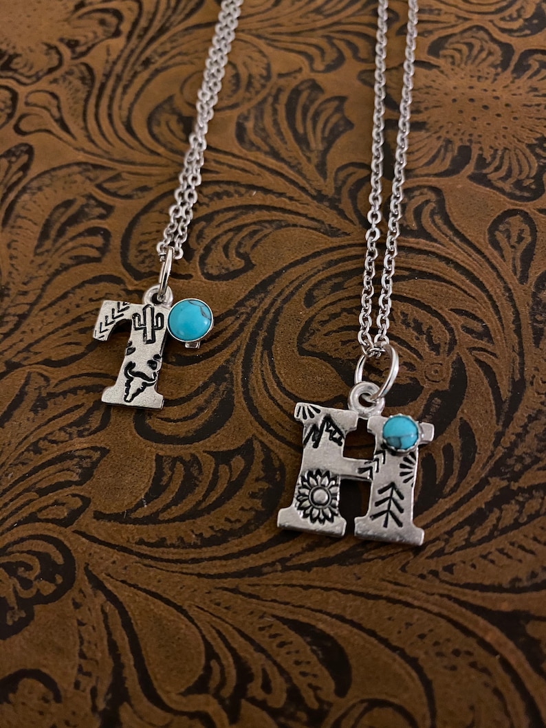 Puede incluir: Dos collares con iniciales plateadas. La letra "T" presenta un dise&ntilde;o de cactus, y la letra "H" tiene un girasol. Ambos colgantes tienen una peque&ntilde;a piedra turquesa y cuelgan de delicadas cadenas. El fondo es una superficie marr&oacute;n texturizada.