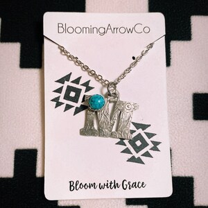 Puede incluir: Un collar plateado con un colgante con la letra "M", adornado con una piedra de color turquesa. El collar cuelga de una tarjeta blanca con los textos "BloomingArrowCo" y "Bloom with Grace", sobre un fondo con un patr&oacute;n en blanco y negro.
