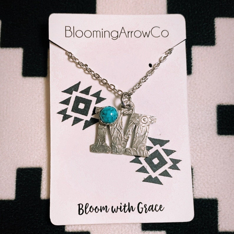 Puede incluir: Un collar plateado con un colgante con la letra "M", adornado con una piedra de color turquesa. El collar cuelga de una tarjeta blanca con los textos "BloomingArrowCo" y "Bloom with Grace", sobre un fondo con un patr&oacute;n en blanco y negro.