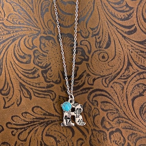 Puede incluir: Collar plateado con un colgante en forma de la letra "R". El colgante presenta una piedra de color turquesa y elementos decorativos. El collar se muestra sobre un fondo marr&oacute;n con un patr&oacute;n floral.