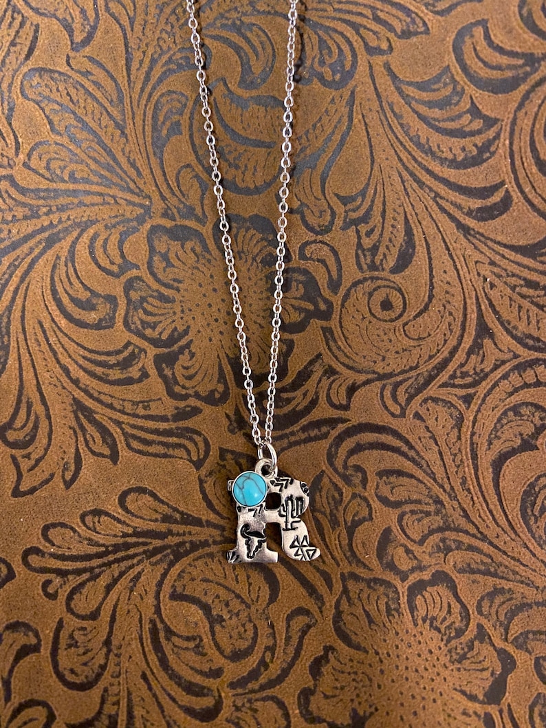 Puede incluir: Collar plateado con un colgante en forma de la letra "R". El colgante presenta una piedra de color turquesa y elementos decorativos. El collar se muestra sobre un fondo marr&oacute;n con un patr&oacute;n floral.