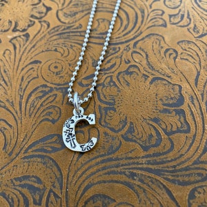 Puede incluir: Un collar plateado con un colgante en forma de la letra "C". El colgante tiene un dise&ntilde;o grabado y cuelga de una delicada cadena de bolas. El fondo es una superficie marr&oacute;n estampada.