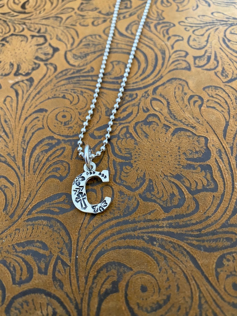 Puede incluir: Un collar plateado con un colgante en forma de la letra "C". El colgante tiene un dise&ntilde;o grabado y cuelga de una delicada cadena de bolas. El fondo es una superficie marr&oacute;n estampada.