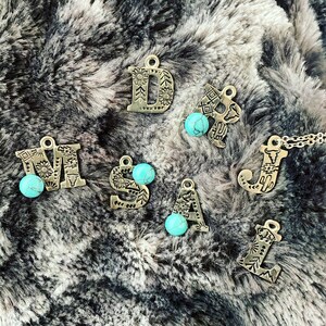 Puede incluir: Amuletos de letras de metal plateado con cuentas de color turquesa. Las letras est&aacute;n decoradas con patrones florales y geom&eacute;tricos. Las letras se muestran sobre una superficie gris y esponjosa.