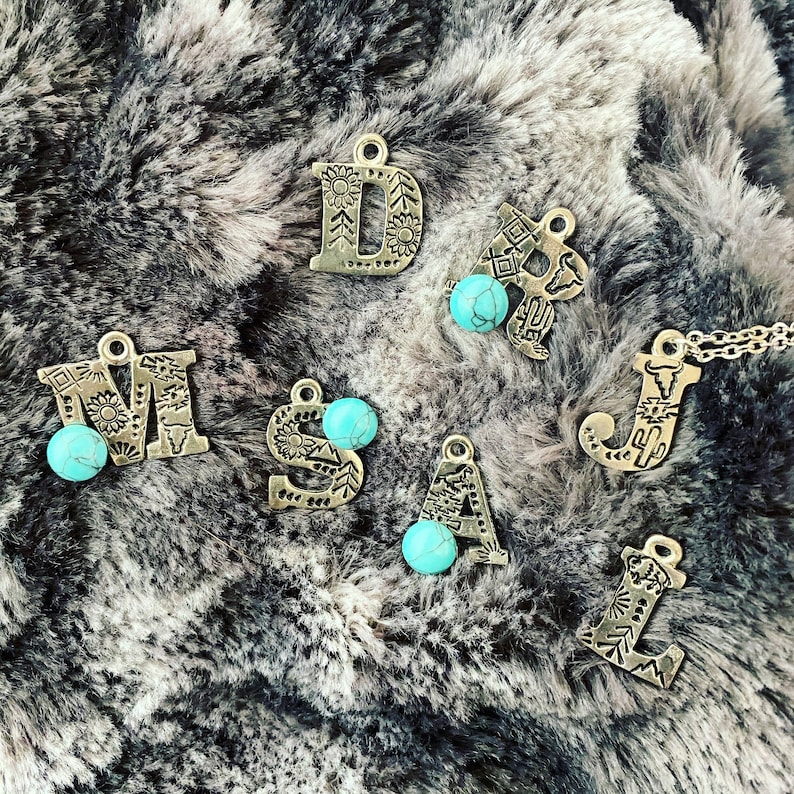 Puede incluir: Amuletos de letras de metal plateado con cuentas de color turquesa. Las letras est&aacute;n decoradas con patrones florales y geom&eacute;tricos. Las letras se muestran sobre una superficie gris y esponjosa.