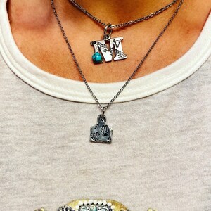 Puede incluir: Dos collares plateados. El collar superior tiene un colgante con forma de letra "M" con una piedra turquesa. El collar inferior tiene un colgante con forma de etiqueta de ganado con un dise&ntilde;o de cactus.