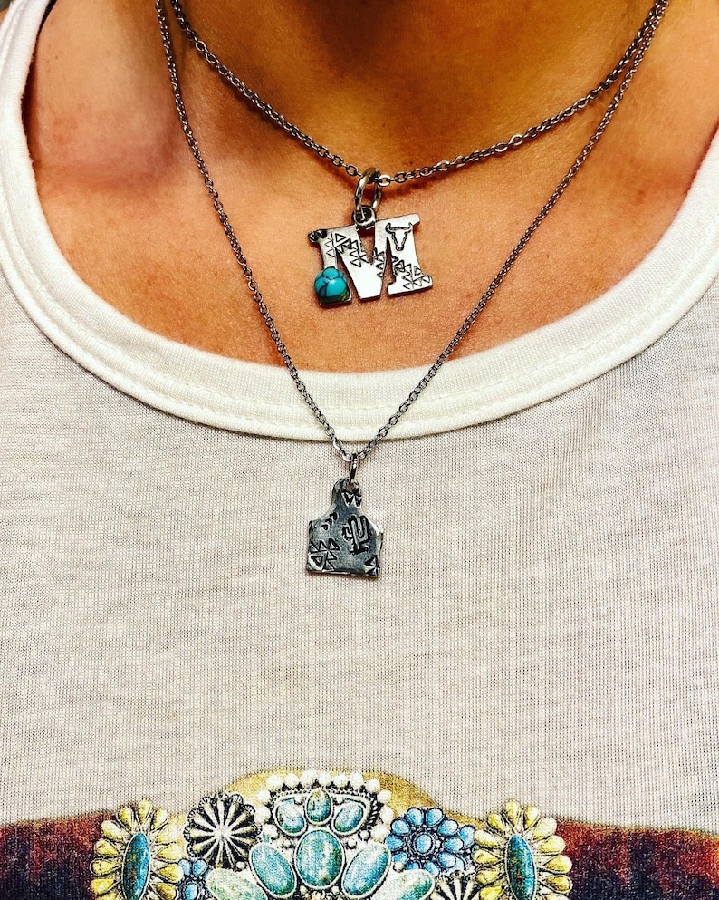 Puede incluir: Dos collares plateados. El collar superior tiene un colgante con forma de letra "M" con una piedra turquesa. El collar inferior tiene un colgante con forma de etiqueta de ganado con un dise&ntilde;o de cactus.