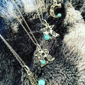 Puede incluir: Collares iniciales plateados con detalles turquesa. Se muestran las letras C, K, R y L, cada una con un dise&ntilde;o decorativo &uacute;nico. Los collares est&aacute;n sobre una superficie texturizada gris y blanca.