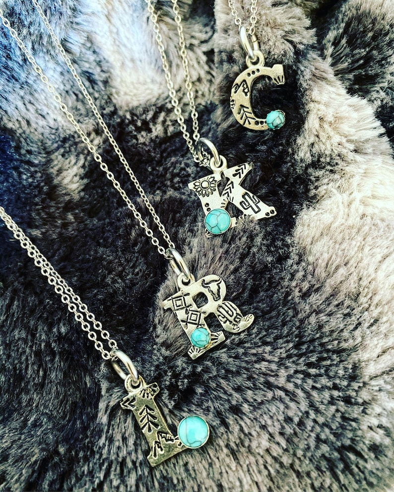 Puede incluir: Collares iniciales plateados con detalles turquesa. Se muestran las letras C, K, R y L, cada una con un dise&ntilde;o decorativo &uacute;nico. Los collares est&aacute;n sobre una superficie texturizada gris y blanca.