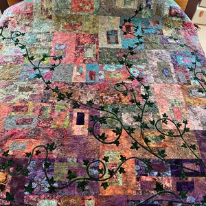 May include: A patchwork quilt in a variety of colours including pink, purple, blue, and green. Dark green vines with leaves are appliquéd across the quilt's surface. The quilt is on a bed, providing a decorative element.