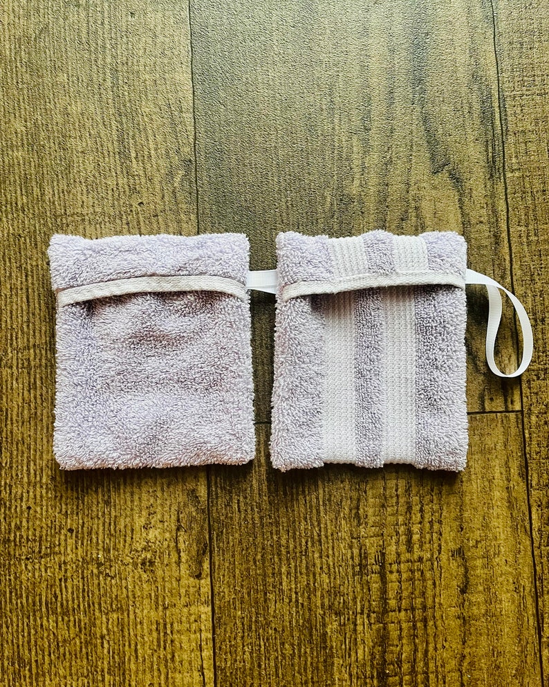 Puede incluir: Dos pa&ntilde;os de lavar de color lavanda con ribete blanco. Un pa&ntilde;o de lavar tiene un patr&oacute;n de rayas y el otro es liso. Ambos pa&ntilde;os de lavar tienen un lazo blanco para colgar.
