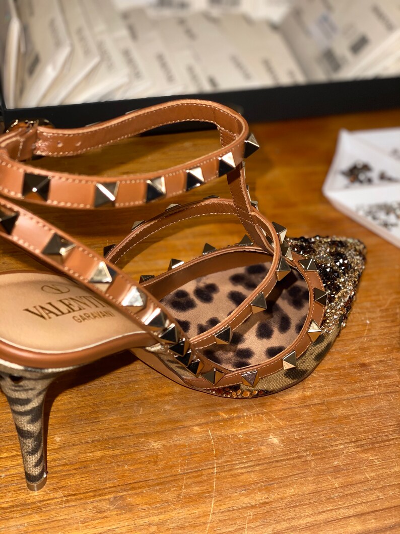 May include: Brown leather high-heeled sandals with gold studs and a leopard print insole. The sandals have a thin ankle strap and a pointed toe. The brand name "VALENTINO GARAVANI" is visible on the insole.