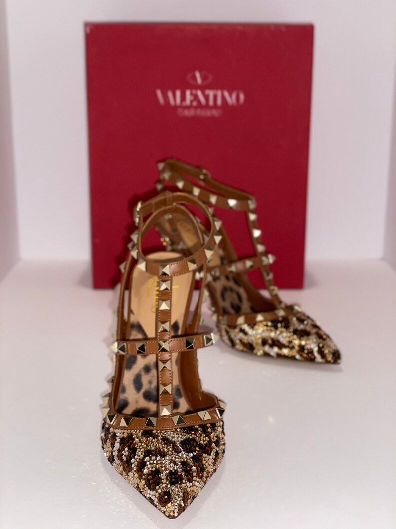 May include: A pair of brown and gold high-heeled shoes with a leopard print design and gold studs. The shoes are on a white surface with a red box behind them. The box has the Valentino Garavani logo on it.