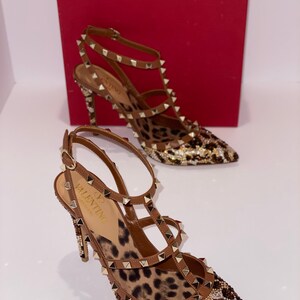May include: A pair of brown leather high-heeled shoes with a leopard print insole and gold studs. The shoes have a pointed toe and ankle straps. The shoes are embellished with rhinestones.