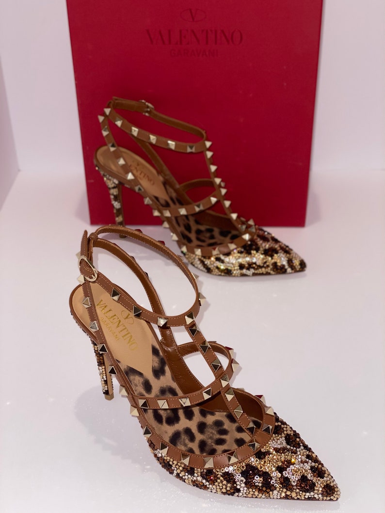 May include: A pair of brown leather high-heeled shoes with a leopard print insole and gold studs. The shoes have a pointed toe and ankle straps. The shoes are embellished with rhinestones.