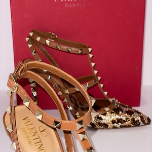 May include: A pair of brown leather high-heeled shoes with gold studs and a leopard print design. The shoes are on a red box with the Valentino Garavani logo.
