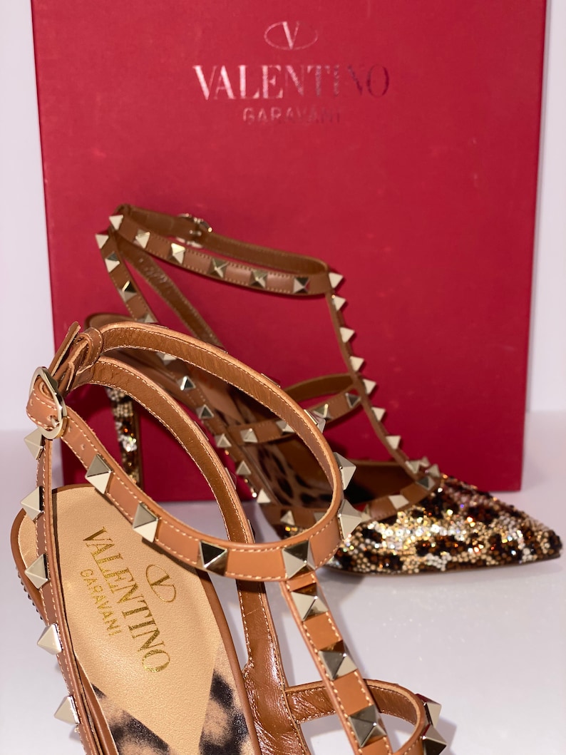 May include: A pair of brown leather high-heeled shoes with gold studs and a leopard print design. The shoes are on a red box with the Valentino Garavani logo.
